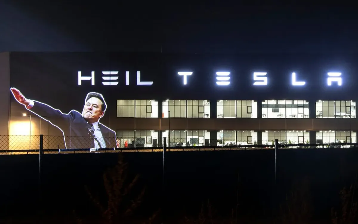 A nighttime photo of a large building with illuminated signage reading 'HEIL TESLA'. Superimposed on the building is a…