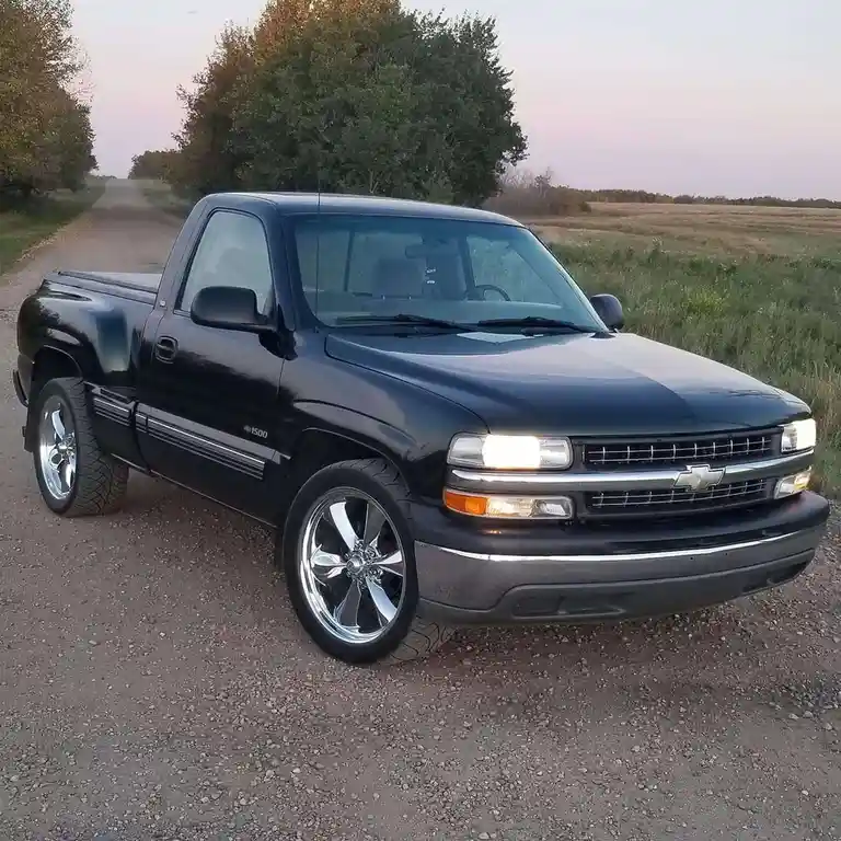 A black Chevrolet 1500 pickup truck with chrome wheels is parked on a gravel road at dusk, surrounded by trees and fields…