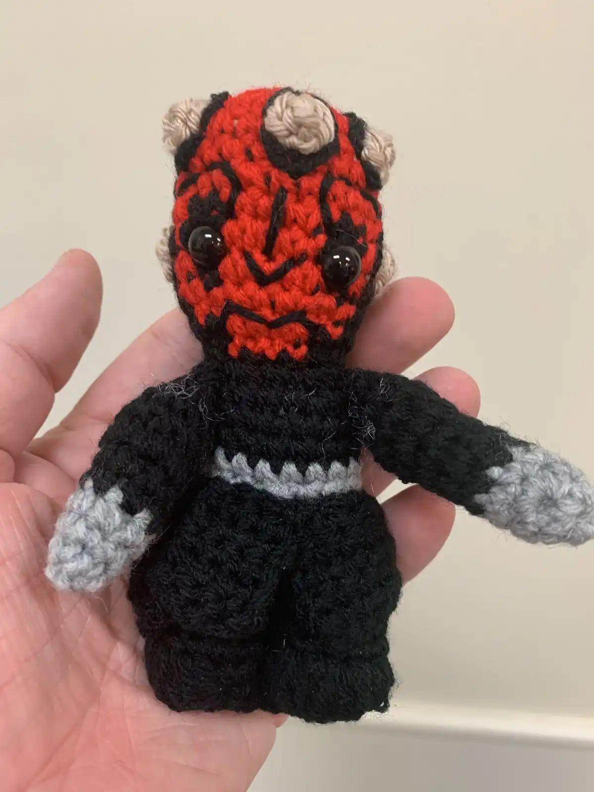 A hand holds a small, crocheted plush toy resembling Darth Maul from Star Wars, featuring red and black yarn for his face…