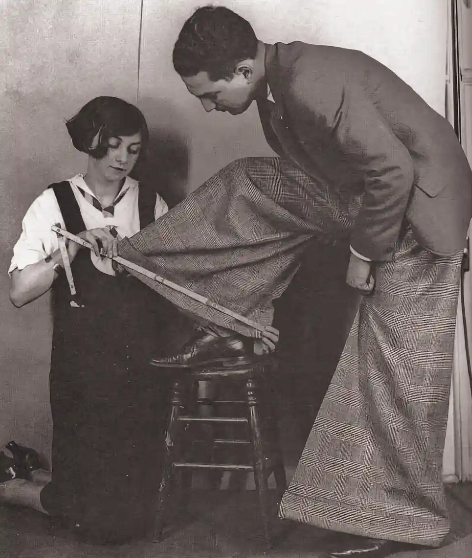 A vintage black-and-white photo shows a woman in an apron measuring a man's leg with a tape measure while he stands on a…