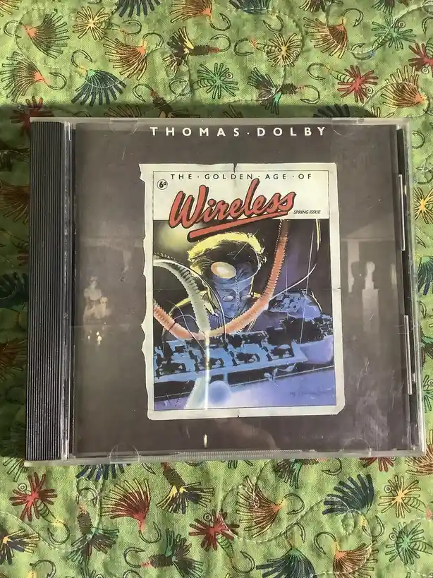 A CD case for Thomas Dolby's album 'The Golden Age of Wireless' is shown, featuring a vintage comic book cover design with a…