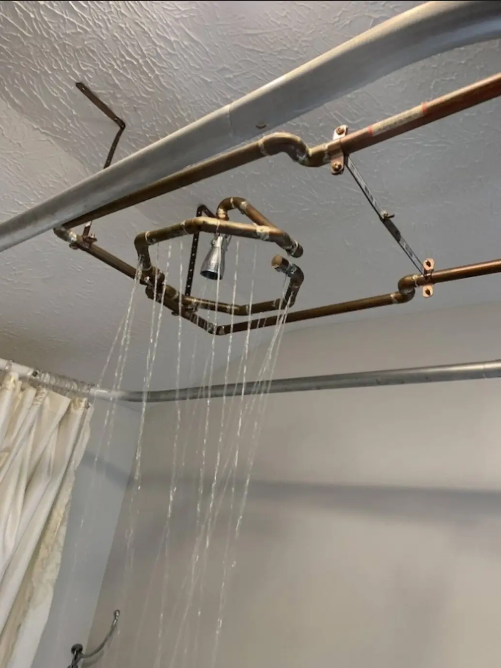 a bathroom ceiling with exposed copper and metal pipes arranged in a complex, angular frame, from which multiple streams of…