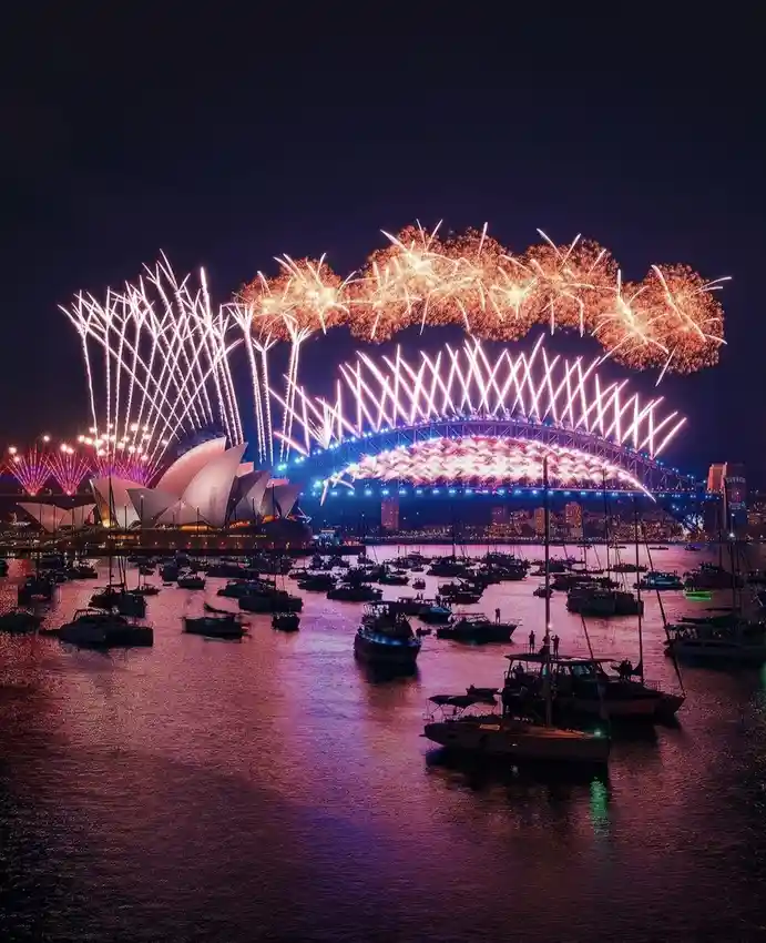 A vibrant nighttime photograph captures a spectacular fireworks display over Sydney Harbour, with the iconic Sydney Opera…
