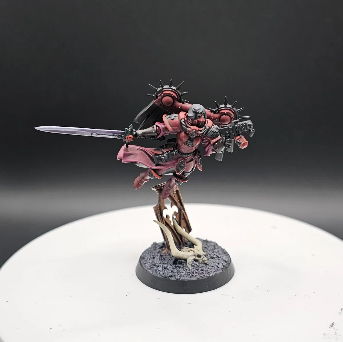 A painted miniature of a red-armored warrior with a sword and spiked shoulder pads stands on a base with a skull and bone…