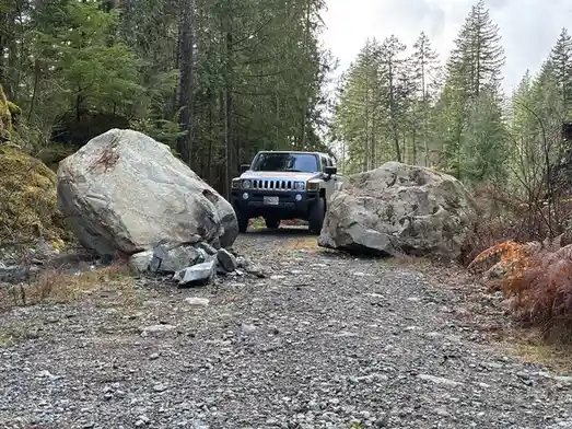 A silver Hummer H2 is parked on a gravel forest road, blocked directly in front of it by two large boulders, one on each…