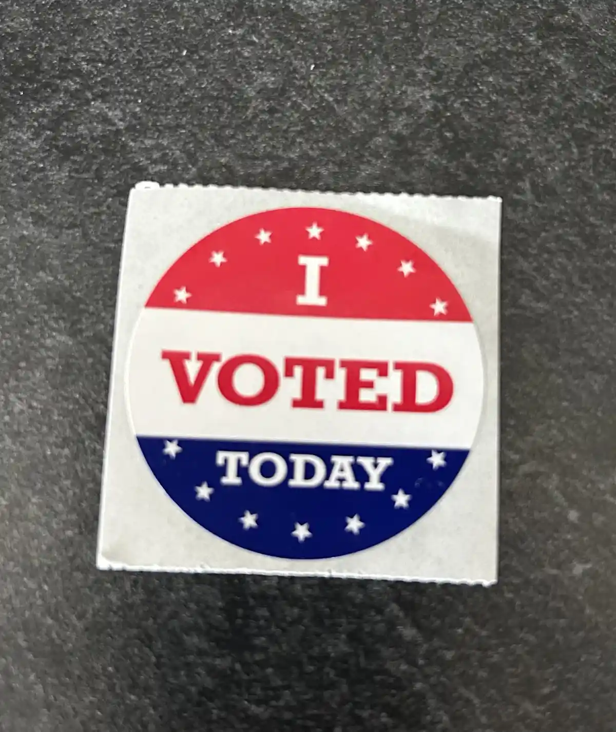 A circular red, white, and blue 'I VOTED TODAY' sticker with white stars on each colored section, resting on a gray textured…