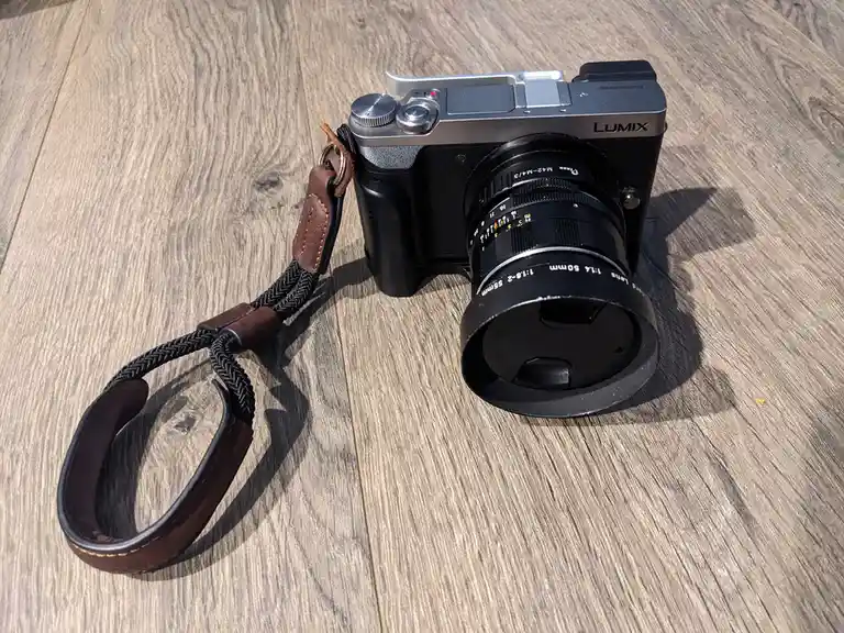 A Panasonic Lumix mirrorless camera with a black lens and brown leather-and-braided strap rests on a wooden surface. The…