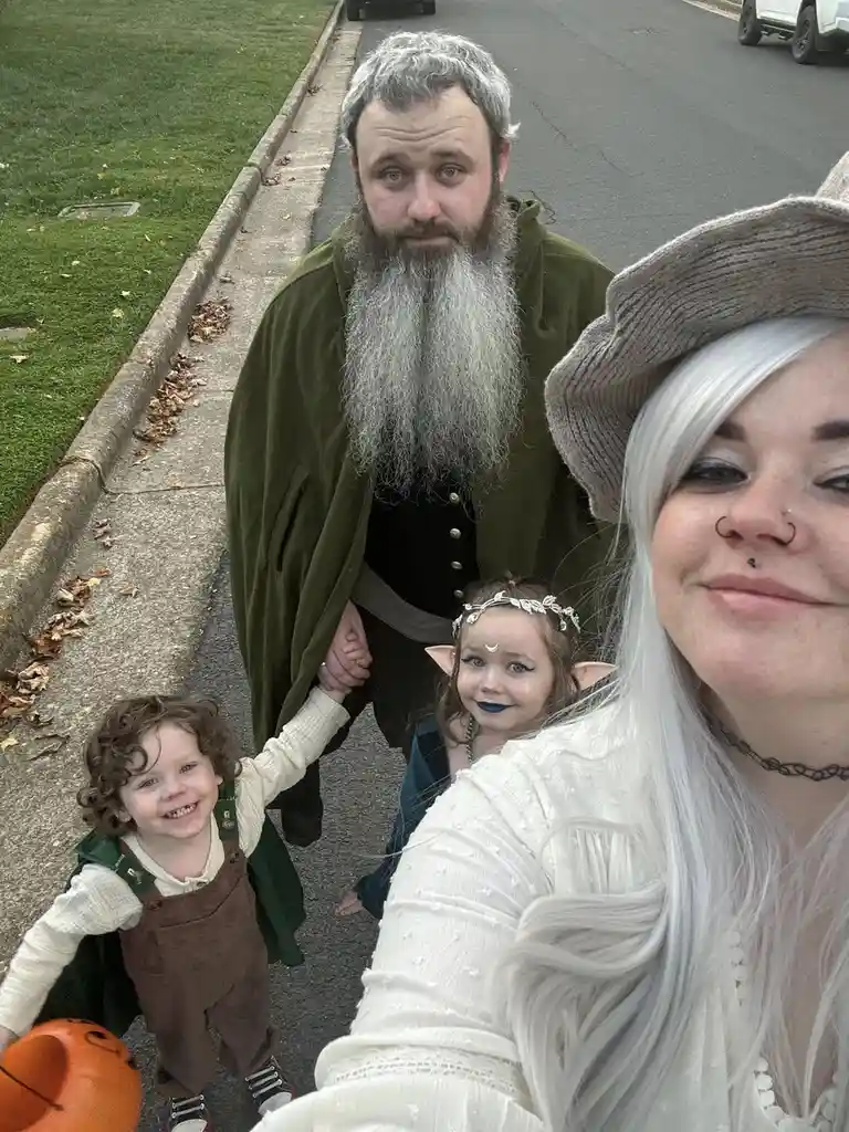 A family of four dressed in fantasy costumes takes a selfie on a residential street. The man in the center has a long gray…