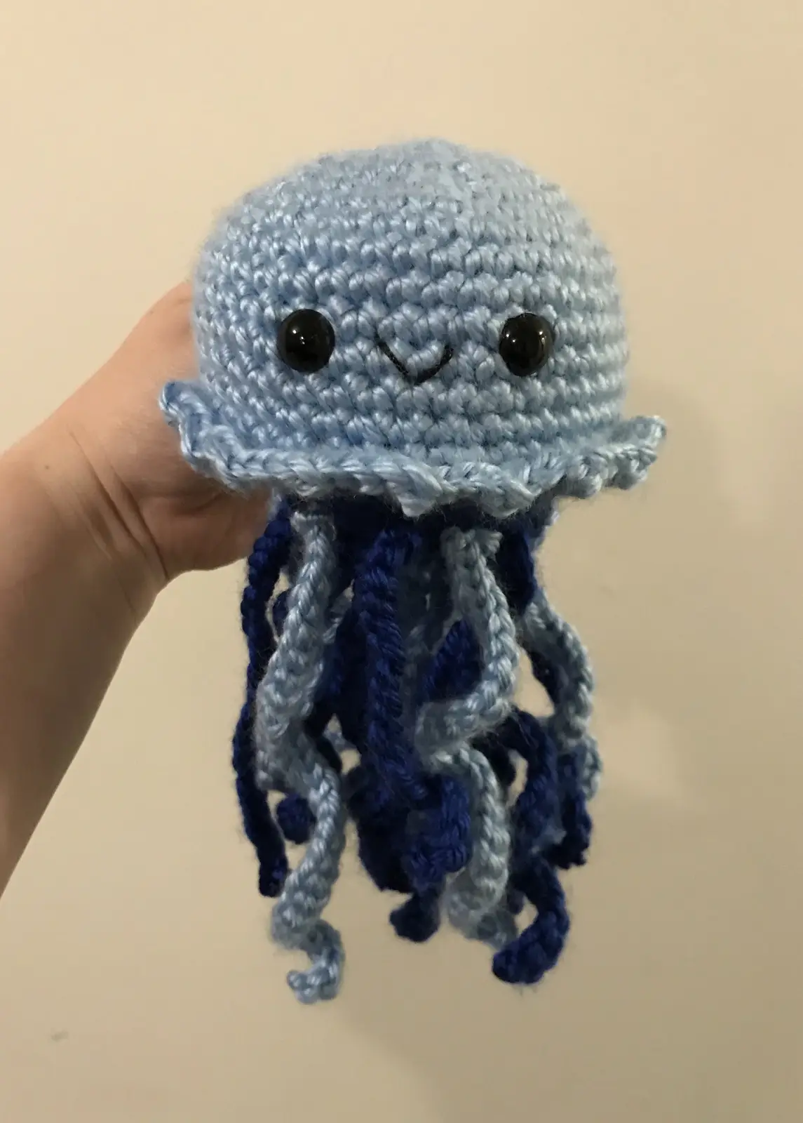 A hand holds a handmade, light blue crocheted jellyfish plushie with large black button eyes and a stitched smile. Its…