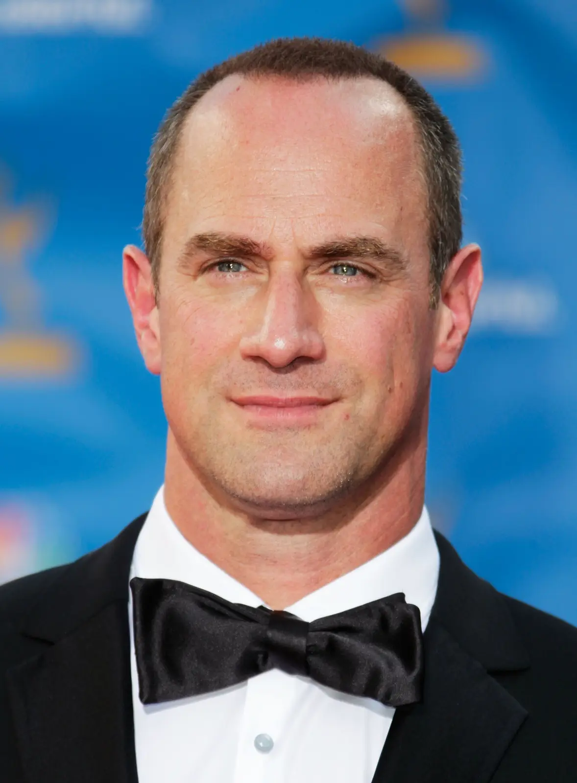 A close-up photo of a man with a receding hairline and short dark hair, wearing a black tuxedo with a bow tie and white…