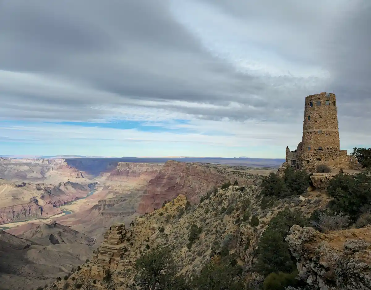 A wide-angle photograph shows the Grand Canyon under a cloudy sky, with a stone watchtower on the right cliff edge…