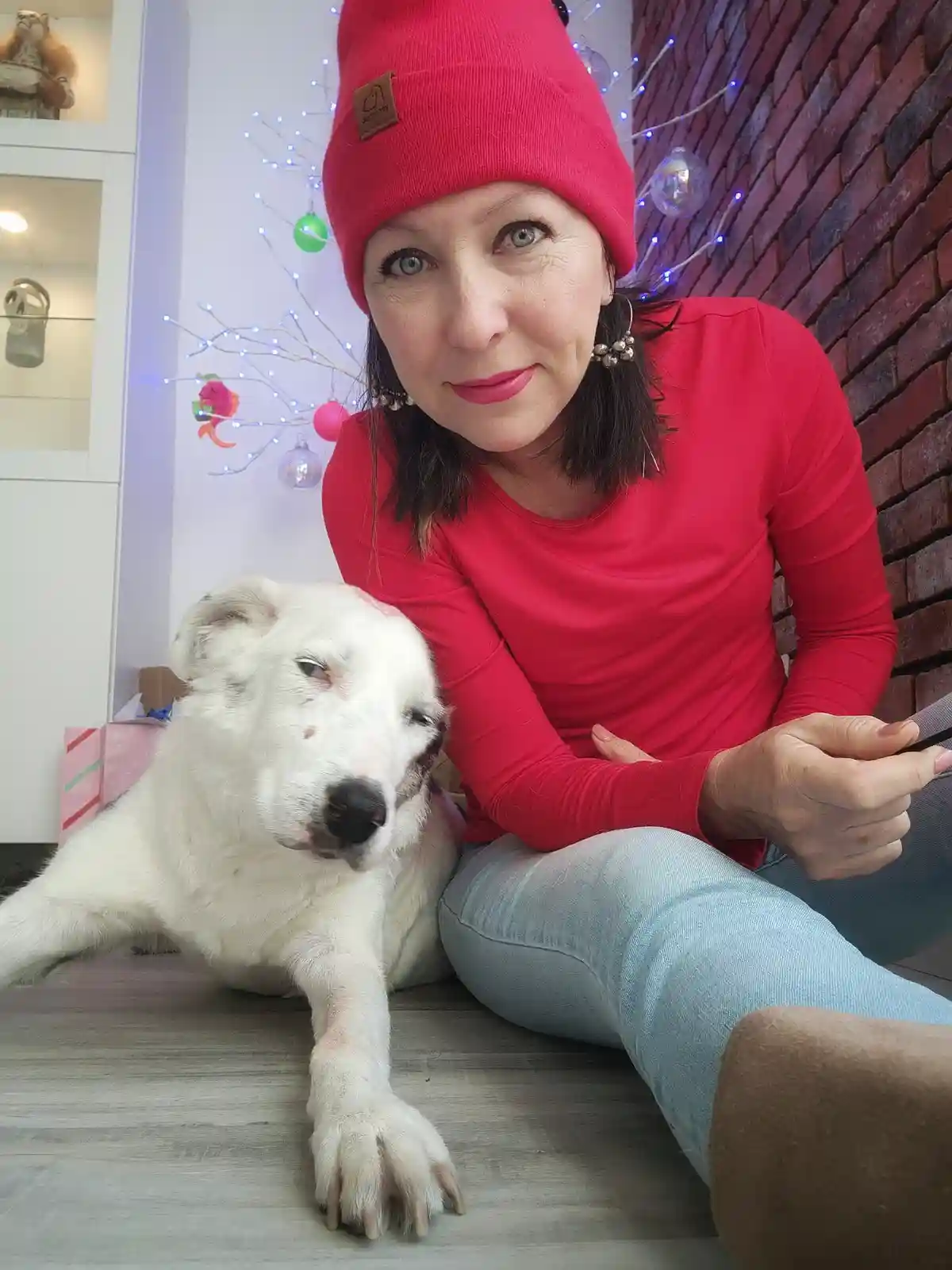 A woman in a red beanie and red shirt sits on a wooden floor next to a white dog with a tired expression. The dog is lying…