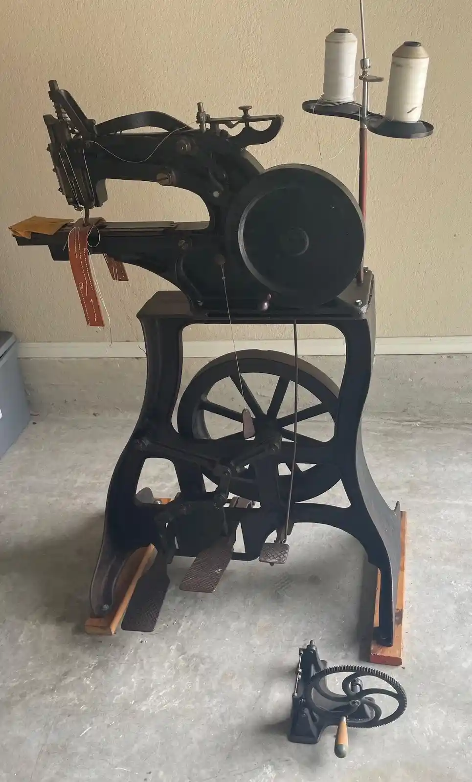 A large, antique black industrial sewing machine with a foot pedal, spools of thread on a stand, and leather straps being…