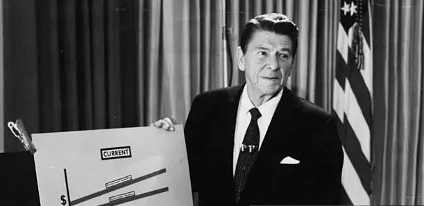 A black-and-white photo of Ronald Reagan in a suit holding a large whiteboard with a graph labeled 'CURRENT' showing a…