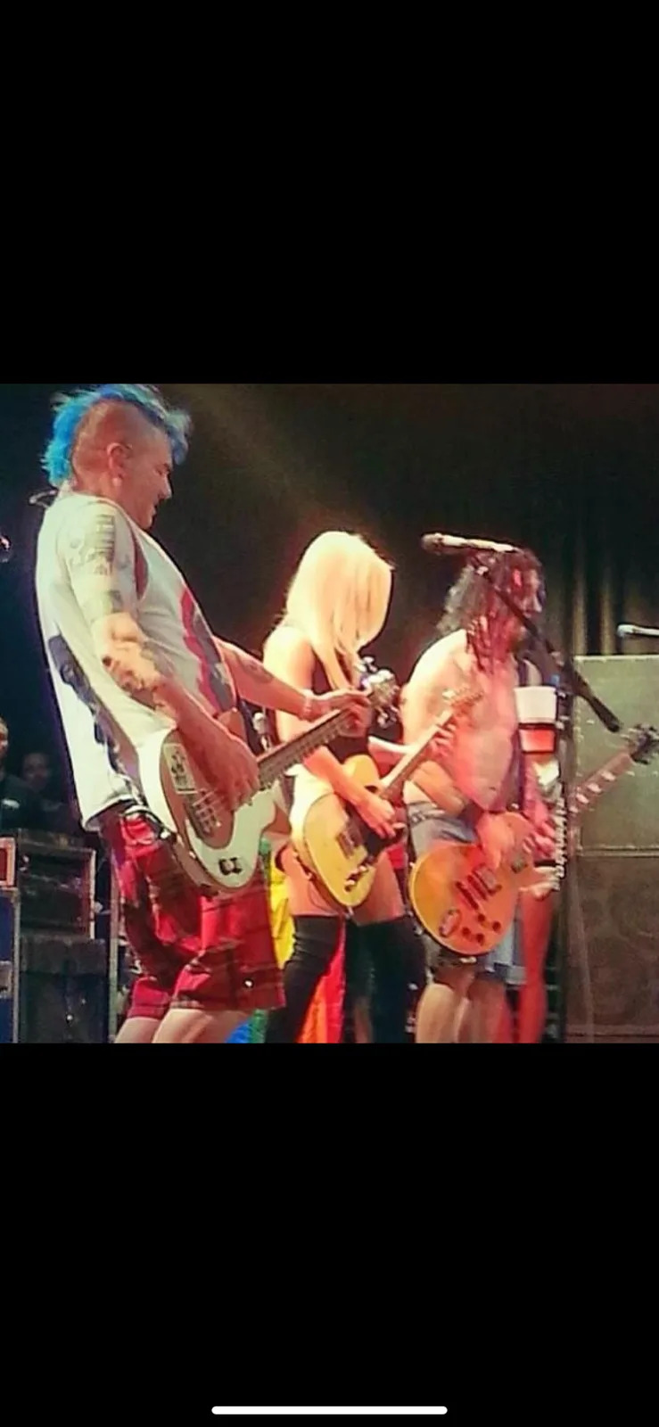Three musicians are on stage during a live concert, each playing electric guitars. The person on the left has blue mohawk…