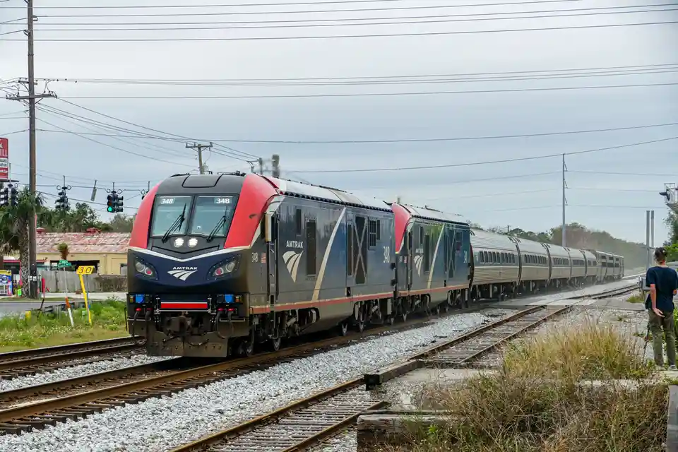 An Amtrak train, numbered 348, is shown moving along railway tracks under an overcast sky. A person stands on the right side…