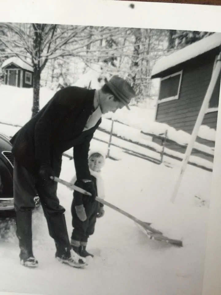 A black-and-white photo shows a man in a suit and hat shoveling snow while a small child in winter clothes stands beside…