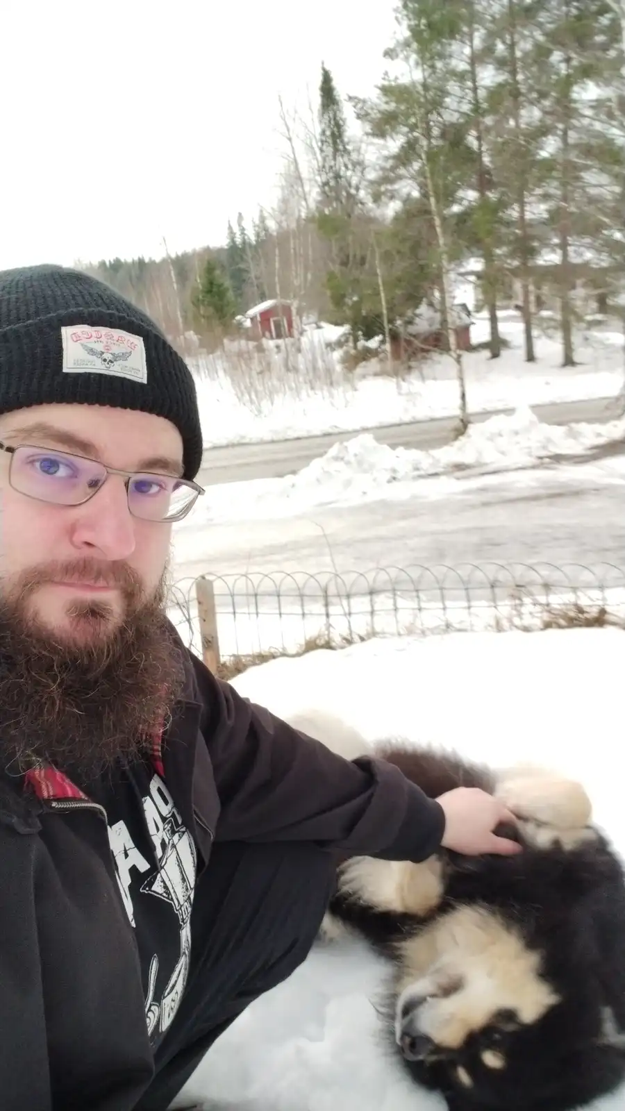 A man with glasses and a beard wearing a black beanie takes a selfie outdoors in snowy terrain while petting a large…