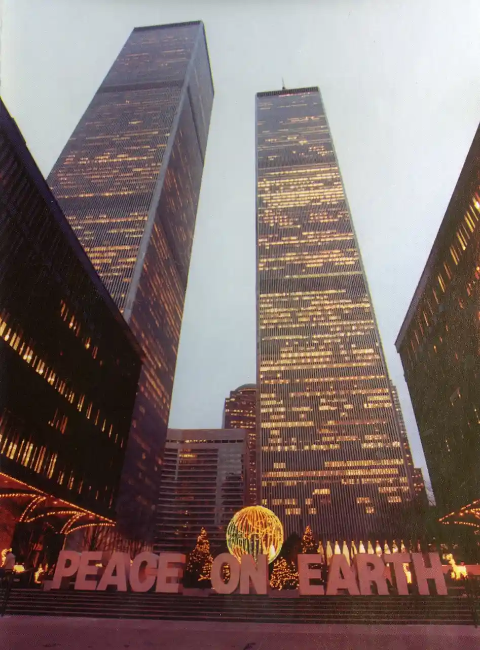 A low-angle photograph shows the Twin Towers of the World Trade Center at dusk, their windows lit up against a pale sky. In…