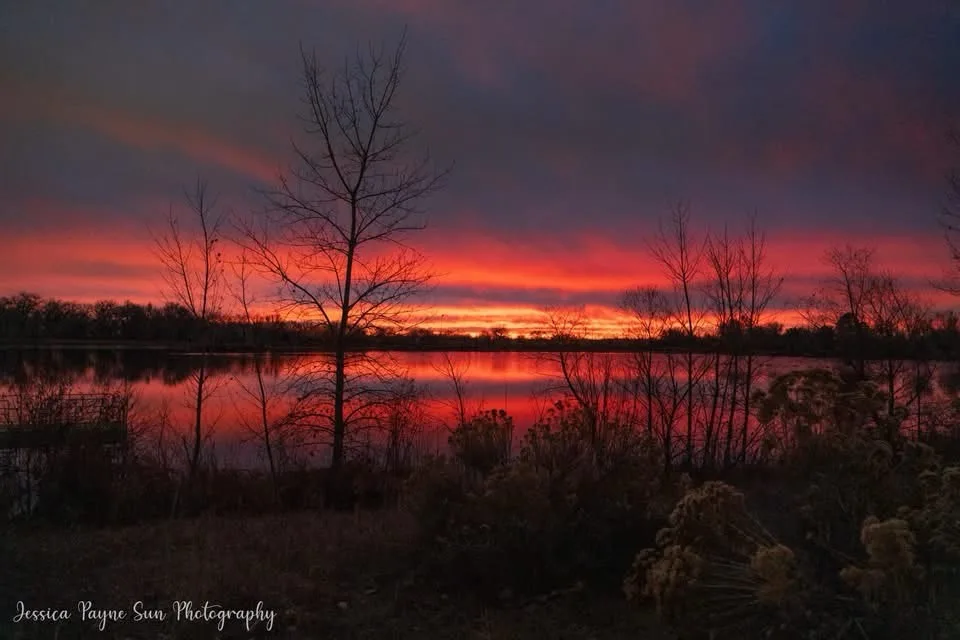 A serene landscape photo of a lake at sunset, with vibrant red and orange clouds reflected in the water, framed by…