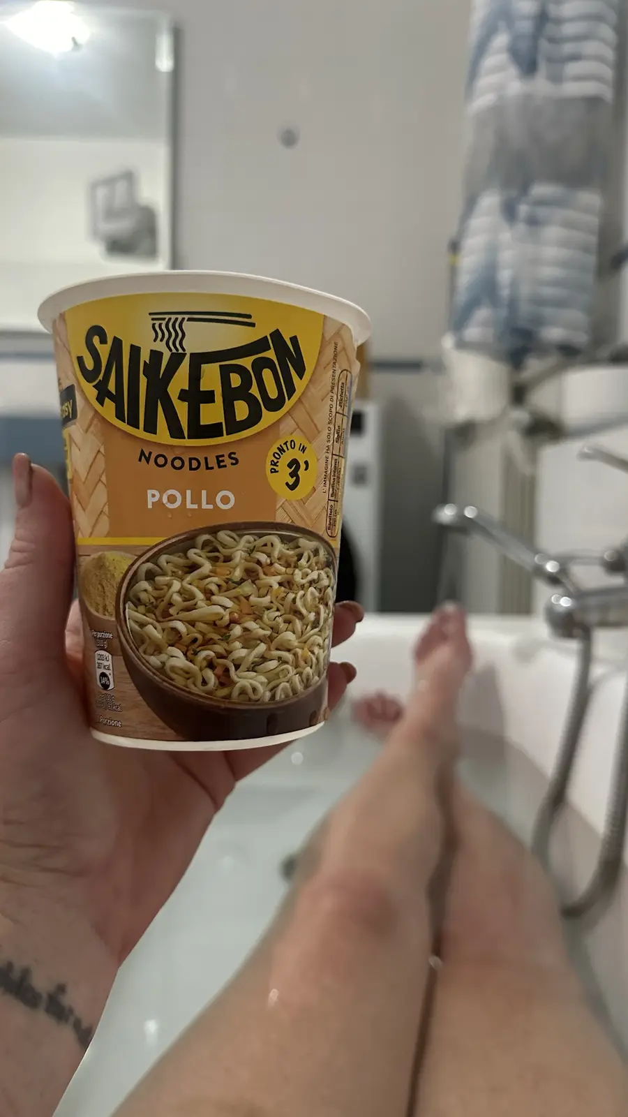 A person is relaxing in a bathtub with their feet up, holding a cup of SAIKEBON noodles labeled 'POLLO' (chicken) and…