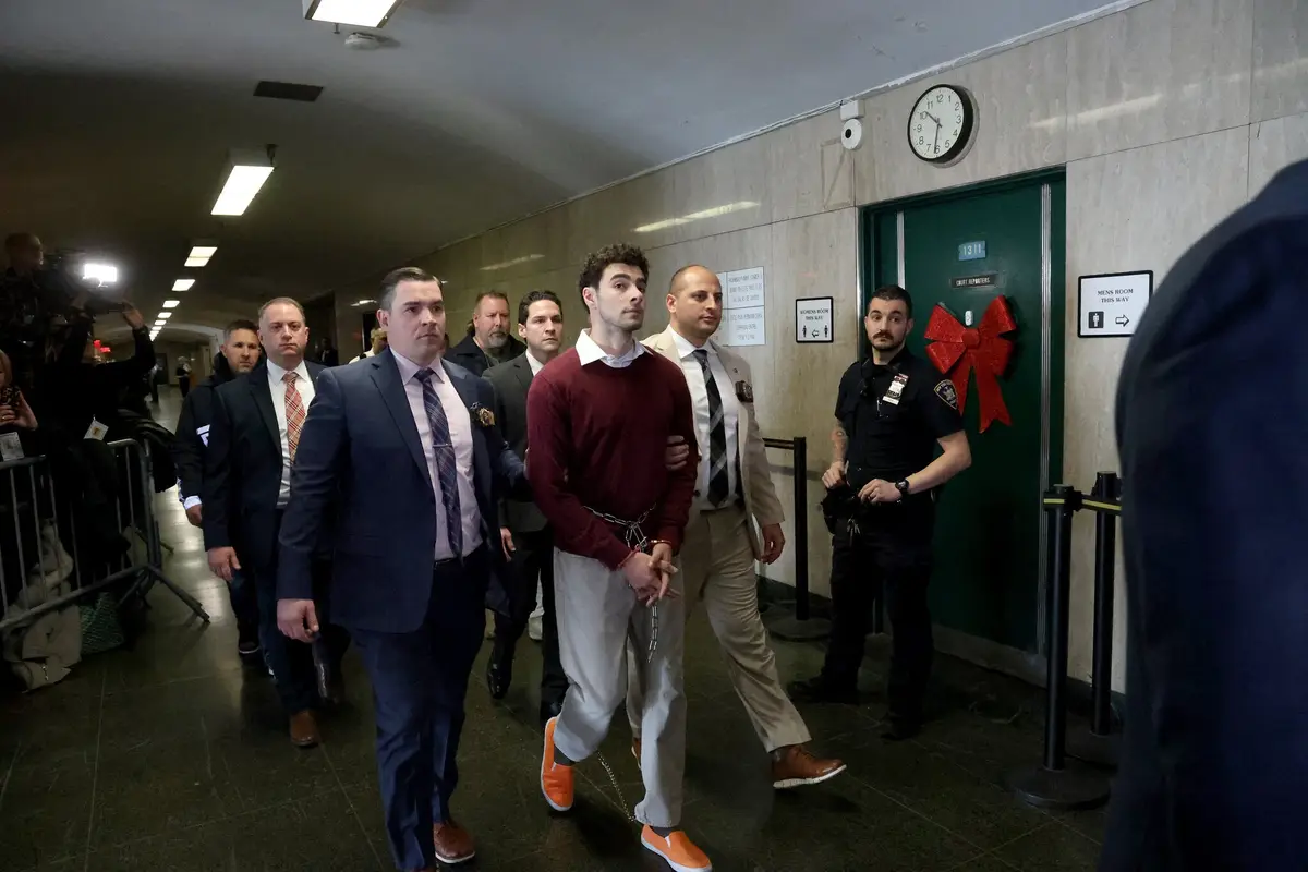 a young man in a maroon sweater and bright orange slip-on shoes is being escorted by law enforcement through a courthouse…