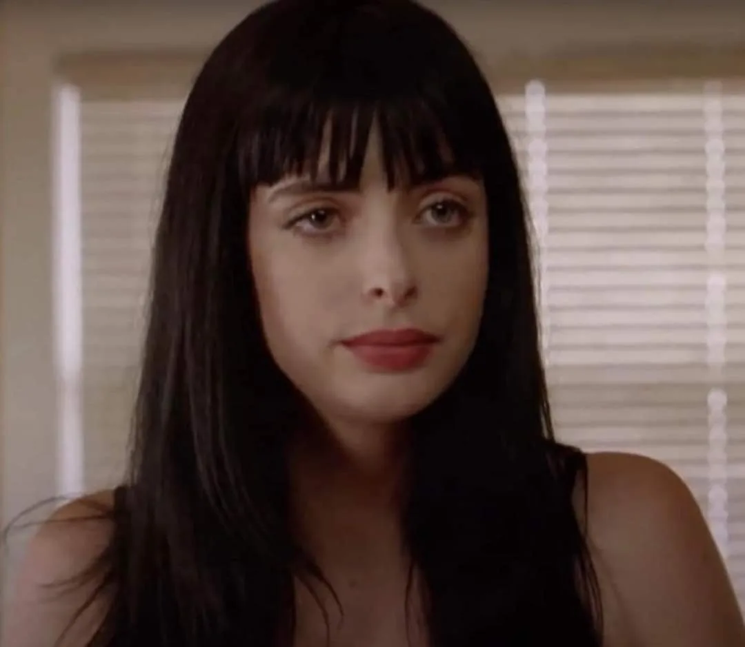 a still image of a woman with long dark hair and bangs, wearing a black top, looking directly at the camera with a calm…