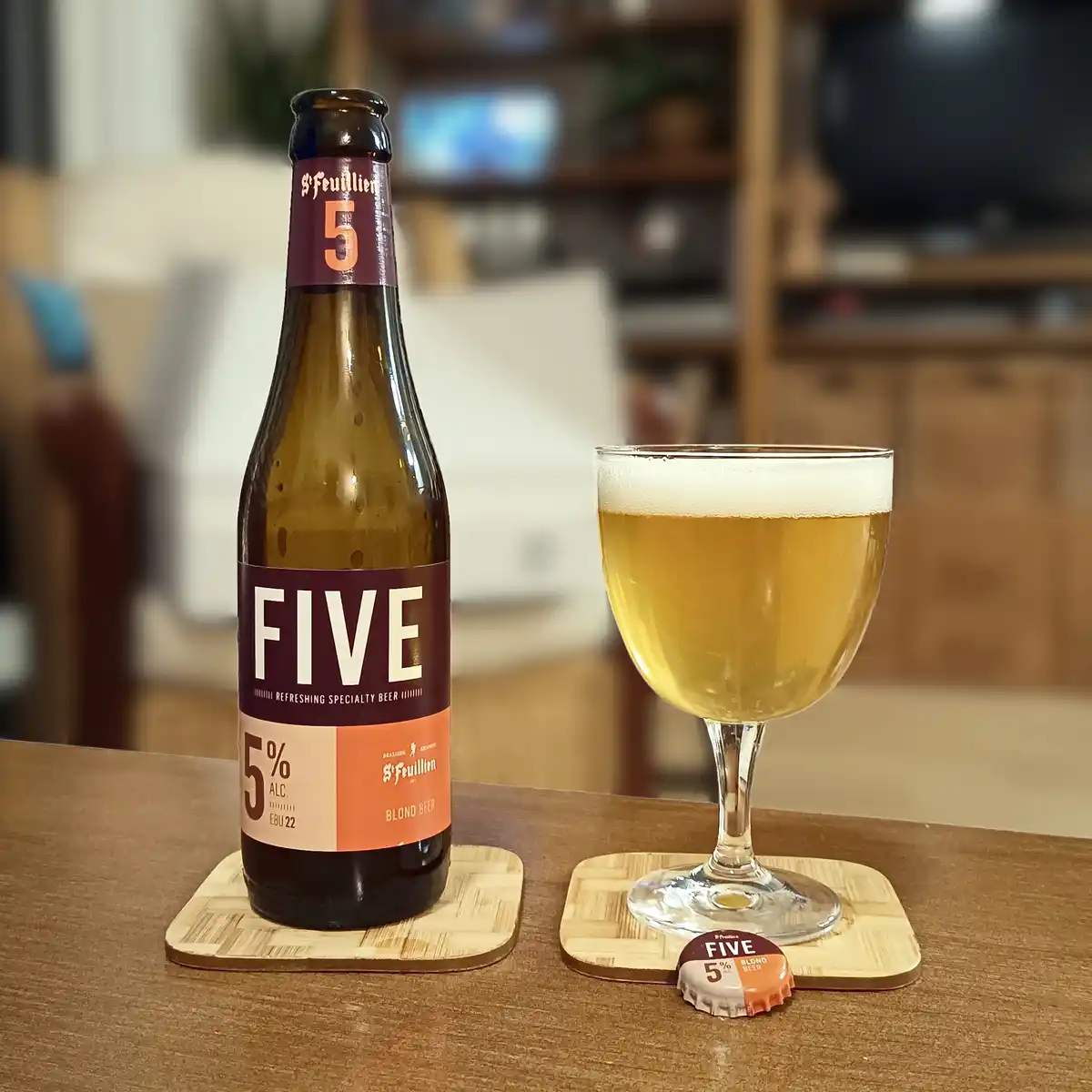 A bottle and glass of St. Feuillien 'Five' blond beer sit on wooden coasters on a table, with the bottle label and glass…