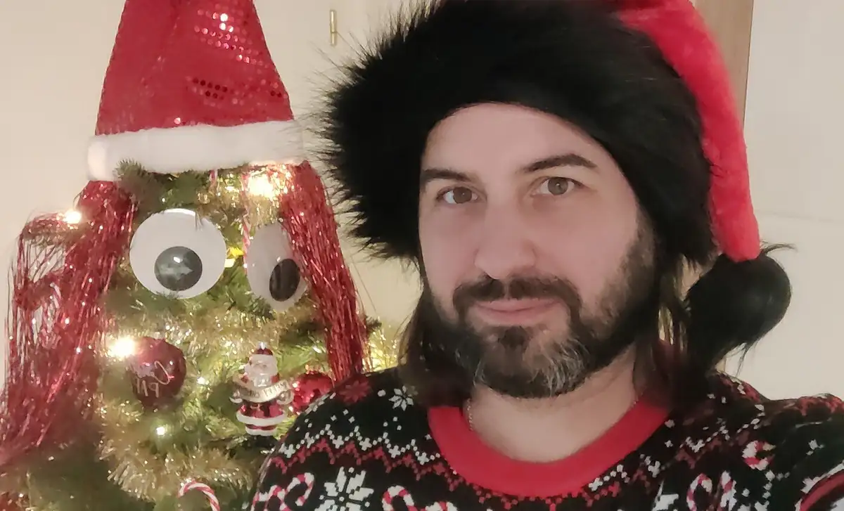 A man with a beard and long hair wearing a red Santa hat and a festive ugly Christmas sweater stands beside a decorated…