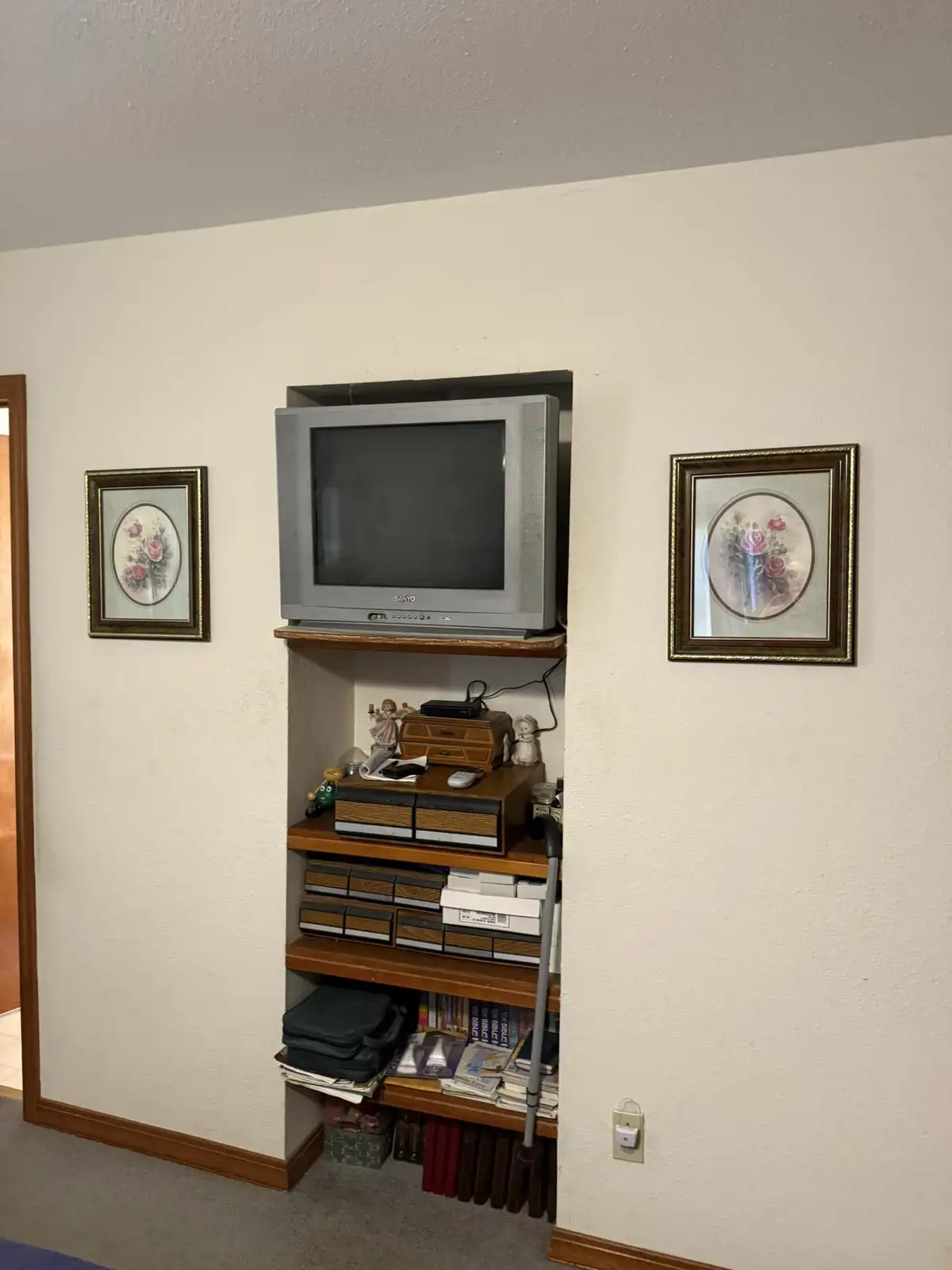 A gray Sanyo CRT television sits on a wooden shelf inside a built-in wall nook, flanked by two framed oval floral pictures…