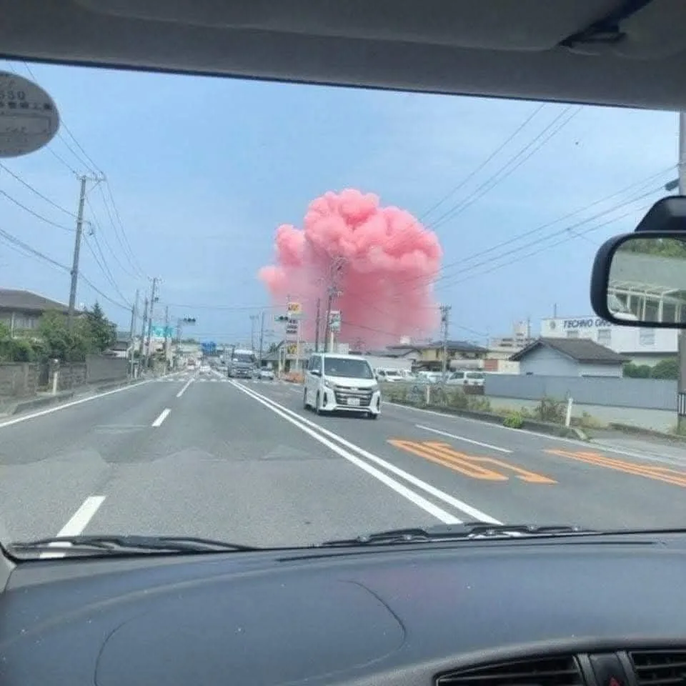 A photo taken from inside a car shows a large, fluffy pink cloud rising in the distance over a suburban street with houses…