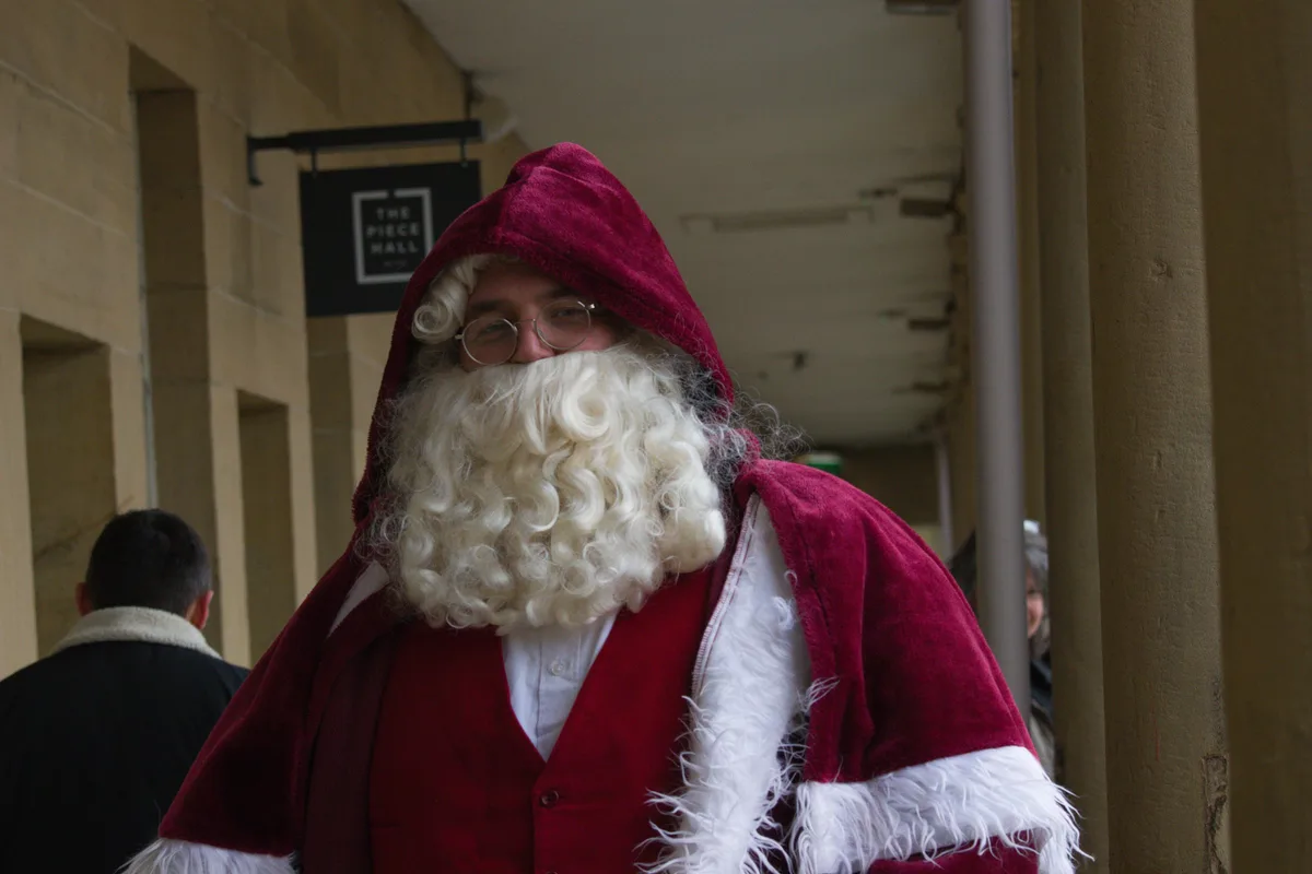 A man dressed as Santa Claus with a large white beard, round glasses, and a red hooded robe with white trim stands in front…
