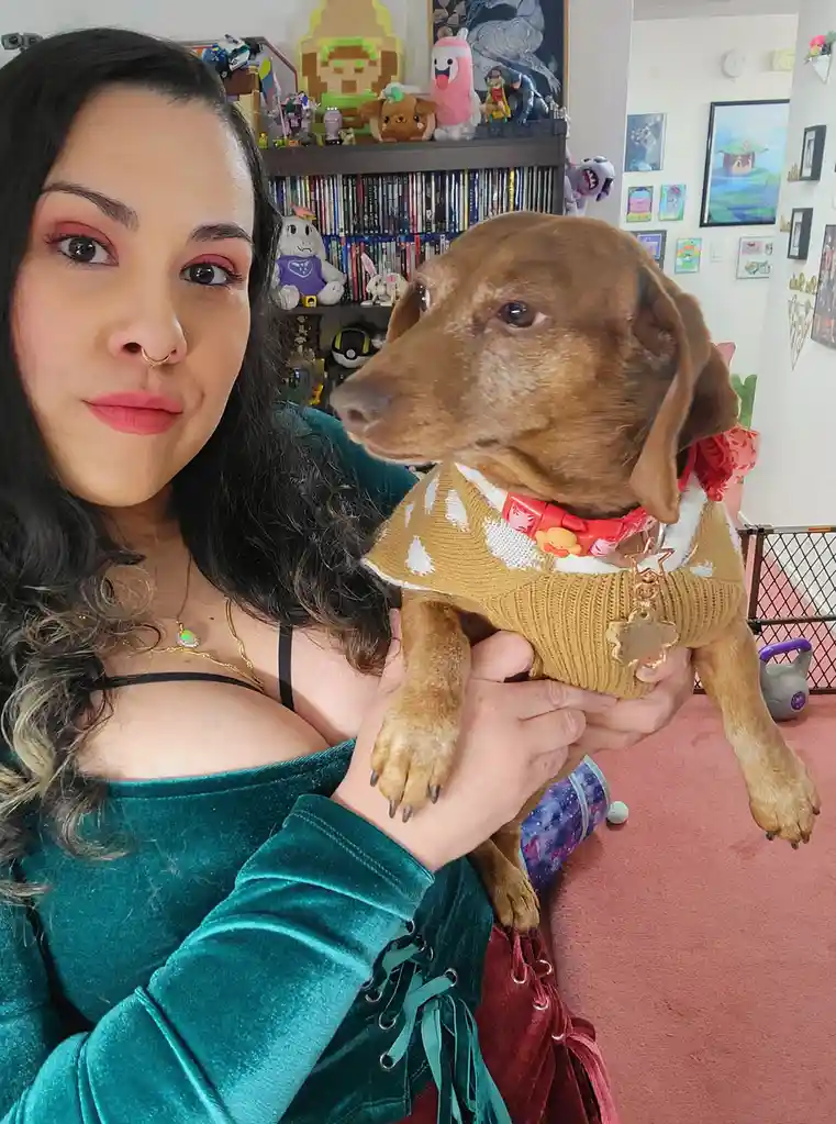 A woman with dark hair and pink eyeshadow holds a brown dachshund wearing a mustard-yellow sweater with white patterns and a…