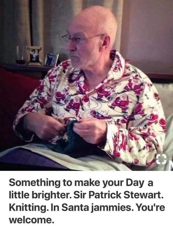 An older man with glasses, wearing pajamas covered in Santa Claus patterns, is seated and knitting a green item. The…