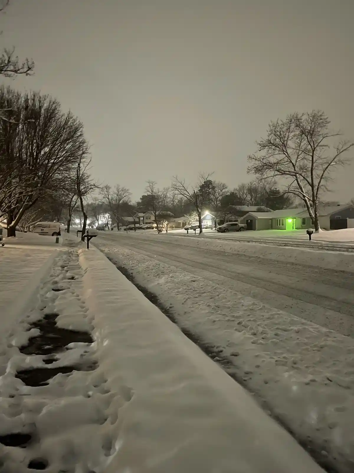 A snowy residential street at night, with snow-covered sidewalks and roads, bare trees, and houses with lights on. One house…