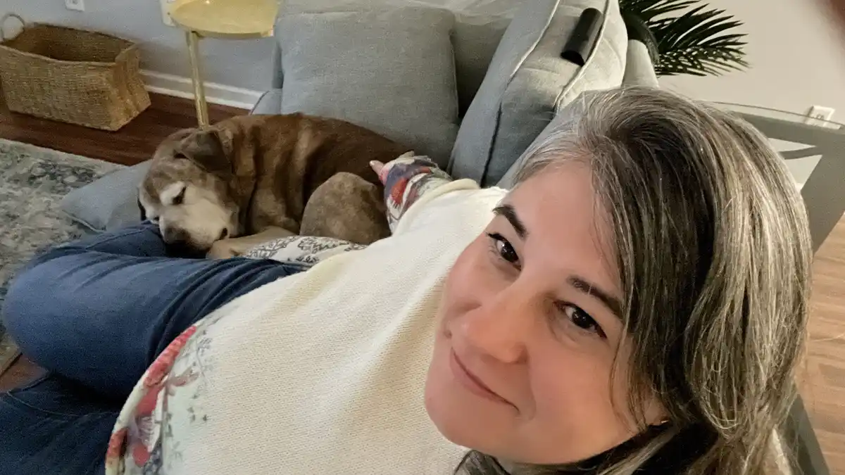 A woman with gray-streaked hair lies on a gray couch, smiling at the camera while a brown and white dog sleeps soundly on…