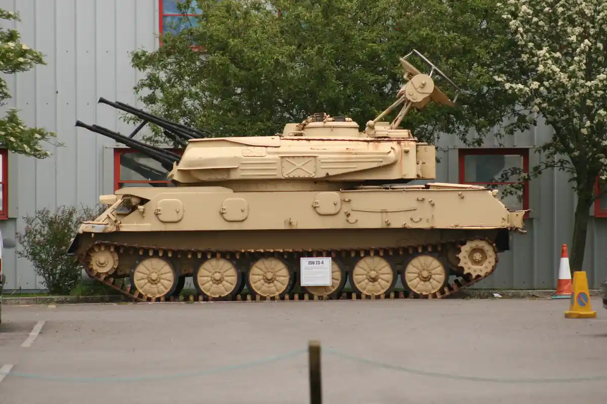 A beige ZSU-23-4 Shilka self-propelled anti-aircraft gun is parked outdoors in front of a gray building with red-framed…