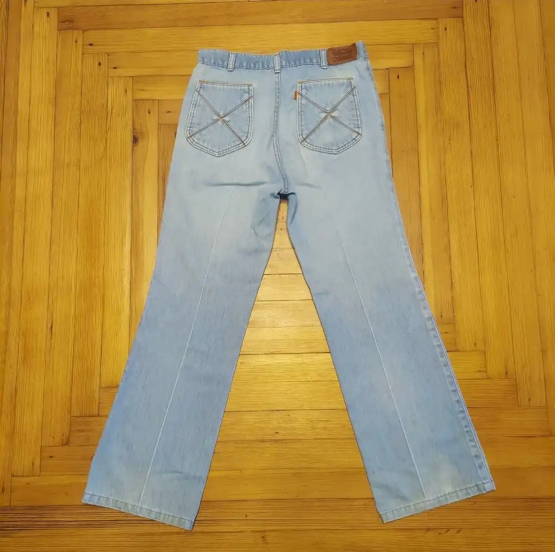 A pair of light blue jeans with distinctive X-stitched back pockets lies flat on a wooden floor, viewed from the rear. The…