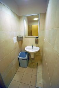 a narrow, beige-tiled bathroom with a small white sink, a mirror above it, a trash can to the left, and a toilet partially…