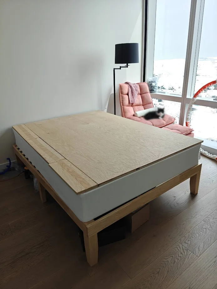 A minimalist bedroom features a bed with a light wood frame and a thick white mattress, but the mattress is completely…