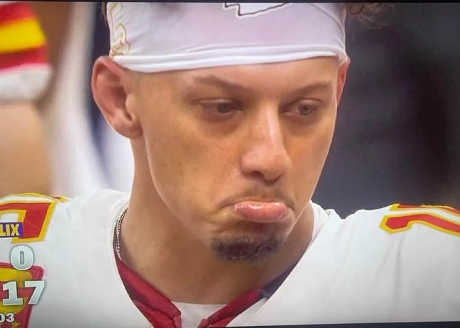 A close-up of a football player wearing a white jersey with red and yellow trim and a white headband, making a pouting face…