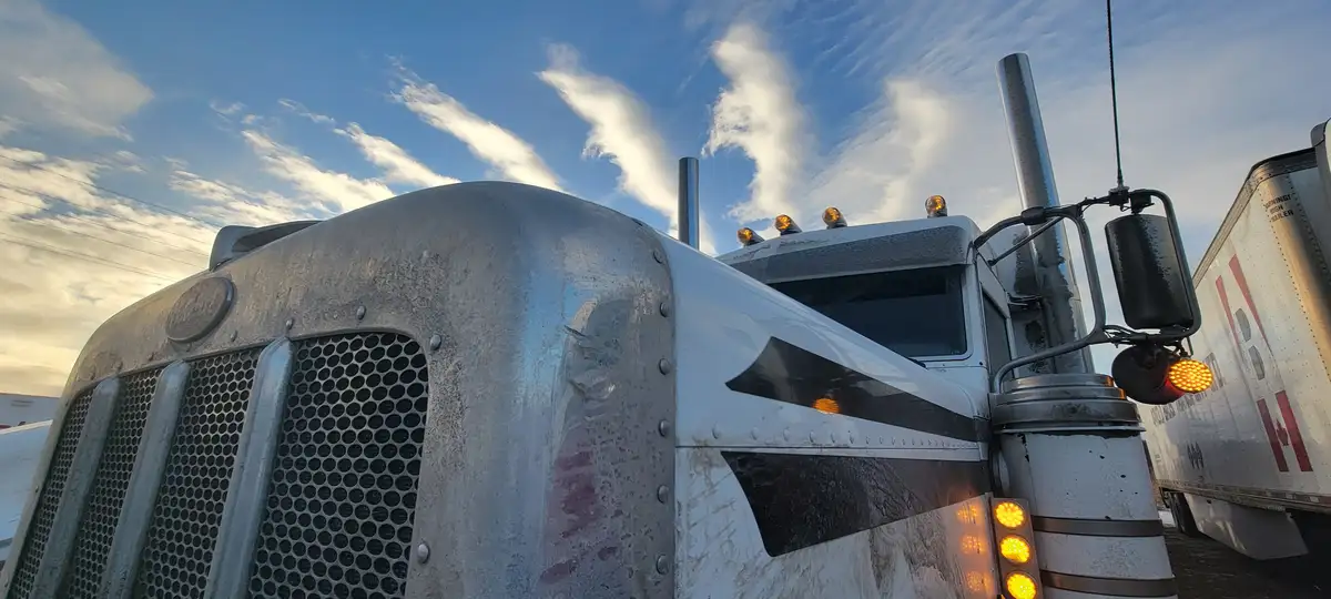 A low-angle photo shows the front of a large, weathered white semi-truck cab with chrome accents, side mirrors, and…