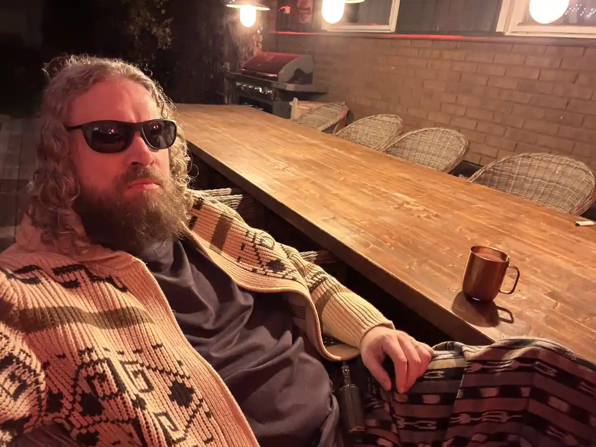 A man with long curly hair and a beard, wearing sunglasses and a patterned knit sweater, sits at a long wooden table…