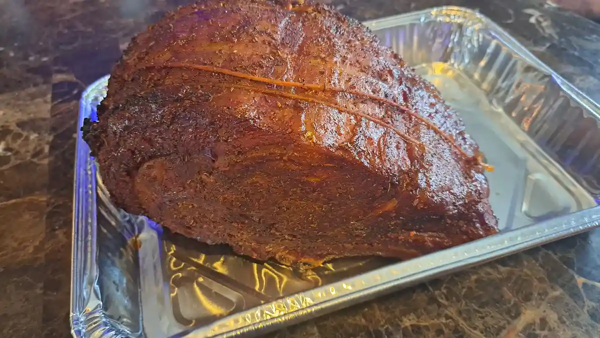 A large, glistening, spice-rubbed roasted meat, likely brisket or pork shoulder, sits in a foil pan on a dark marble…