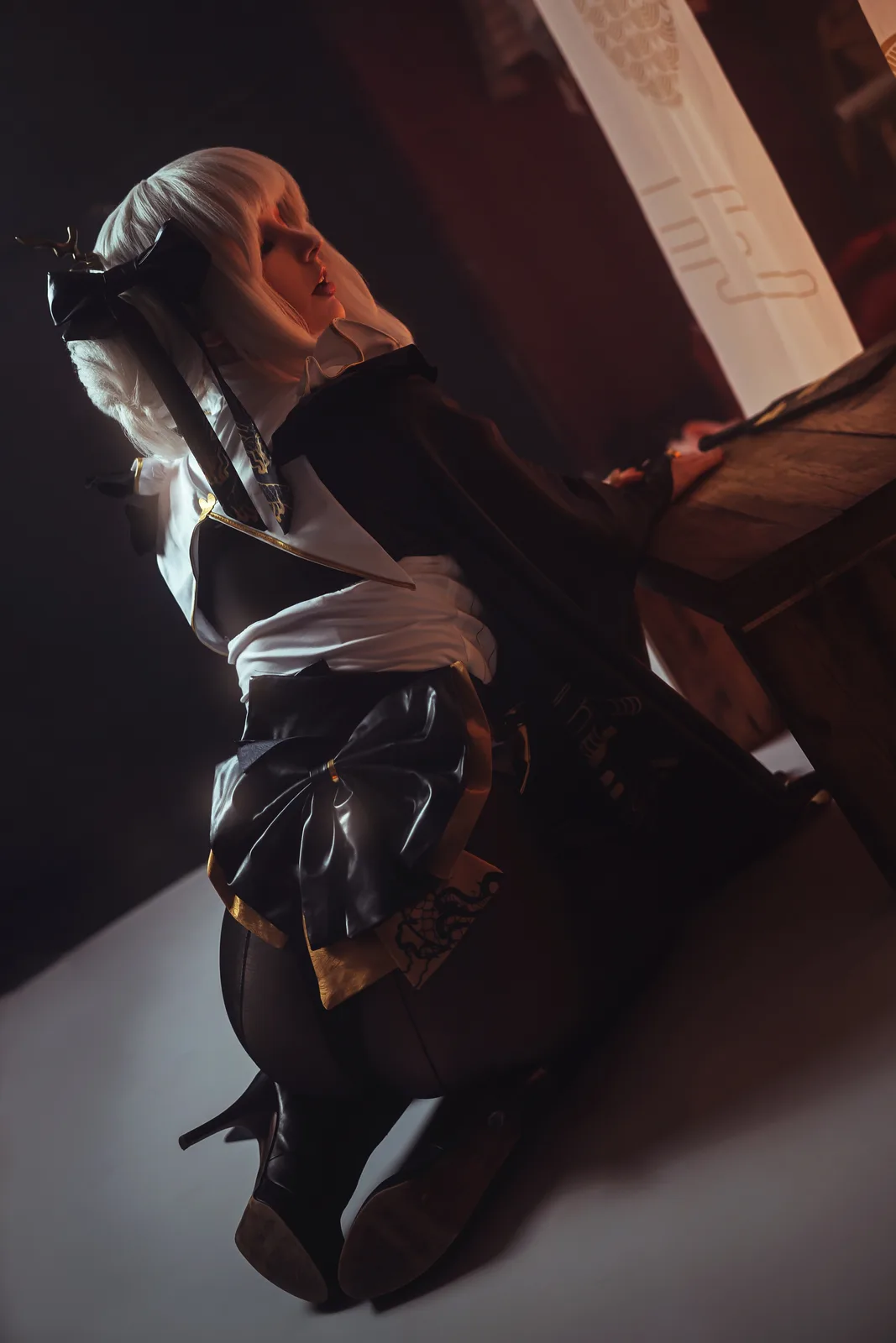 A person in a detailed cosplay outfit with white hair, black bow, and gold trim leans back against a wooden table in a dimly…