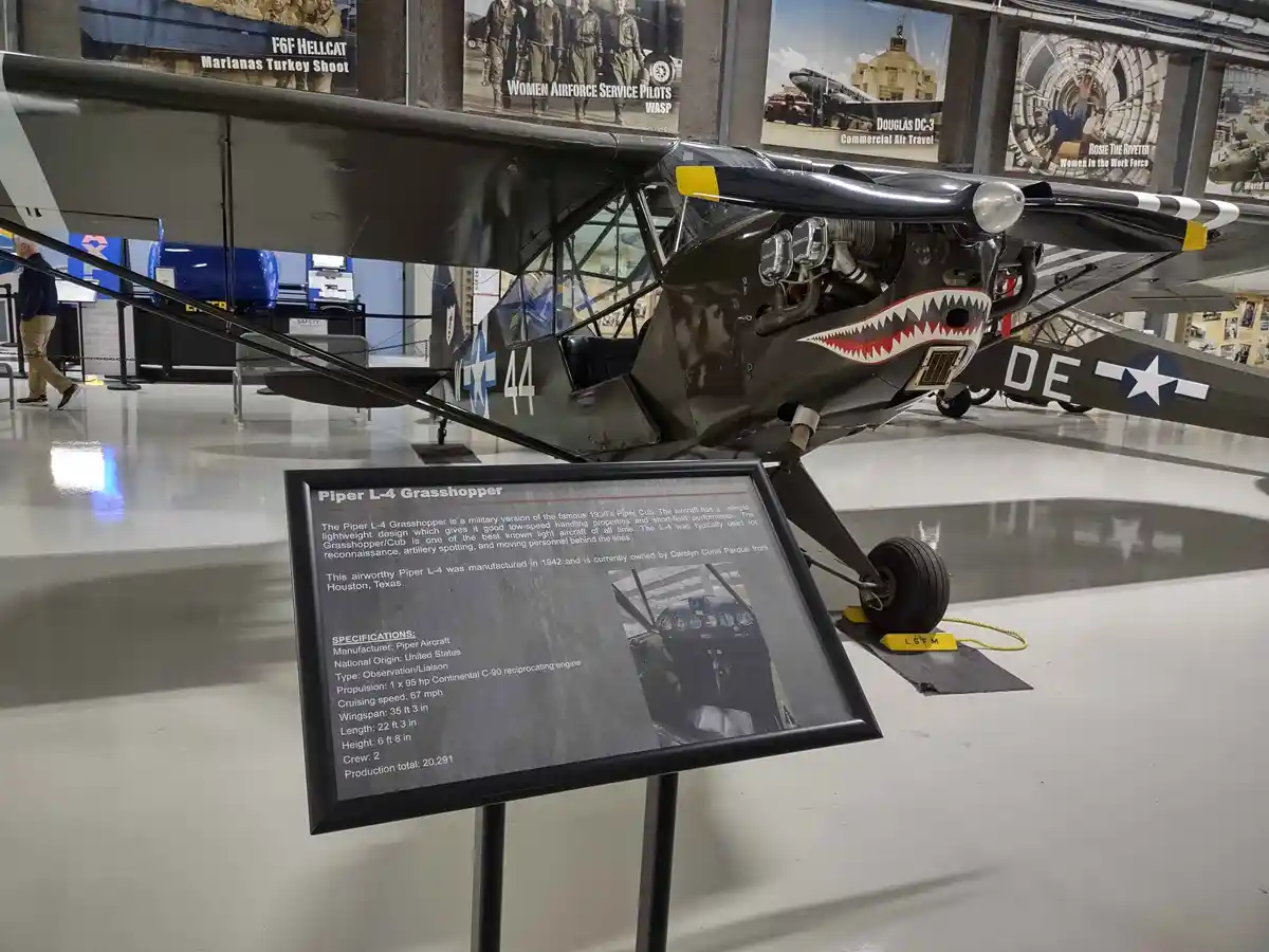 A black Piper L-4 Grasshopper aircraft with a shark mouth painted on its nose is displayed in an aviation museum. The plane…