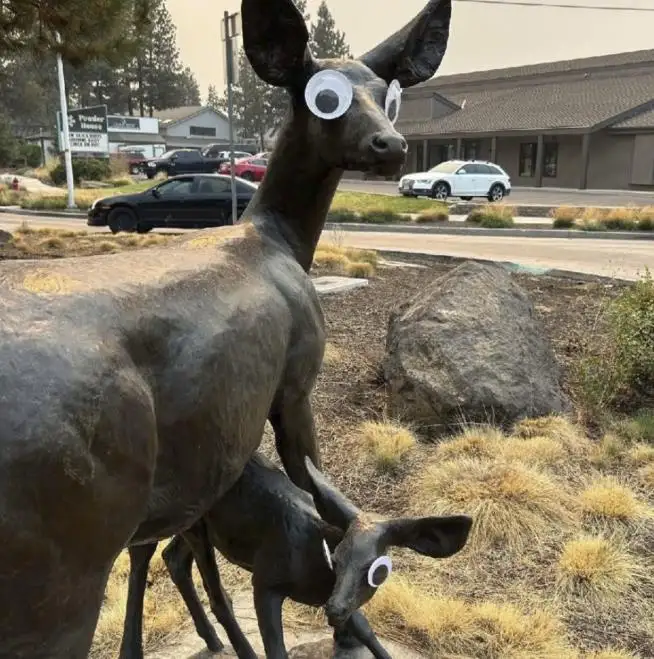 A bronze statue of a mother deer and her fawn, both with large, cartoonish googly eyes pasted onto their heads, standing in…