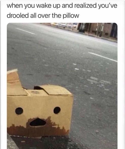 A cardboard box cut into a simplistic face with two round eye holes and a triangular mouth sits on a wet asphalt road. The…