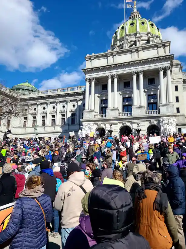 A large crowd of protesters wearing winter coats and holding signs gathers in front of the Pennsylvania State Capitol…