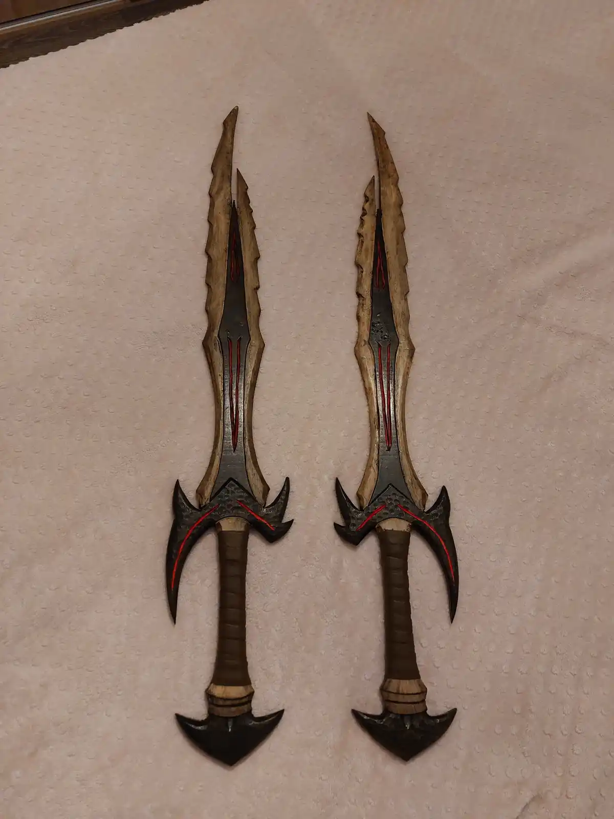 Two intricately crafted fantasy-style swords with jagged blades, red accents, and dark hilts lie side by side on a textured…